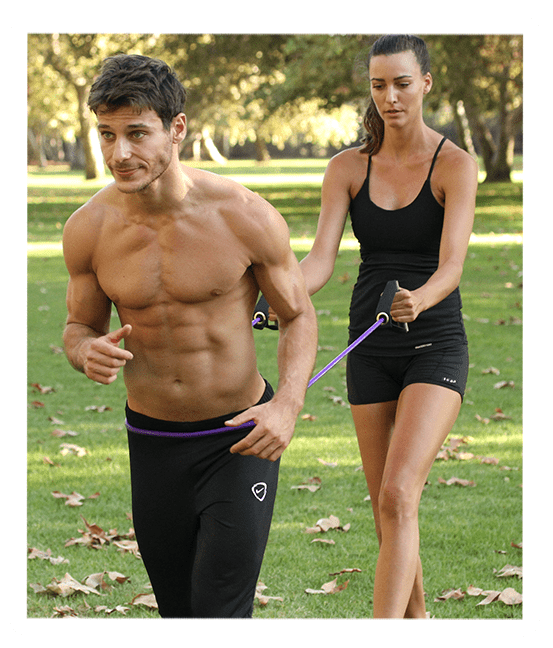 A shirtless man runs forward in a grassy park, pulling a purple resistance band held by a woman behind him.